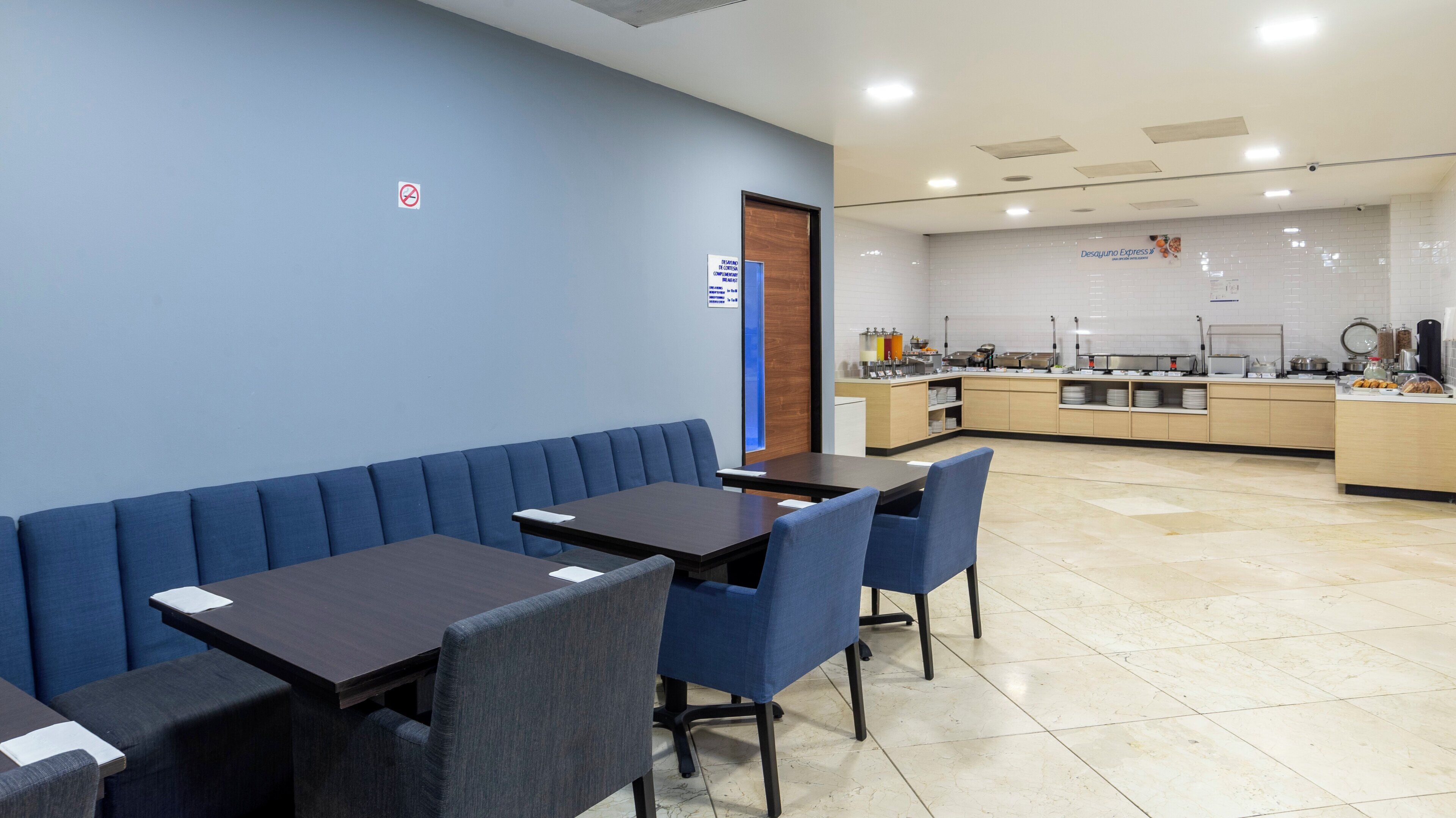 Un restaurant moderne avec des banquettes et des tables bleues, un espace cuisine et une enseigne indiquant « Desayuno Express ».