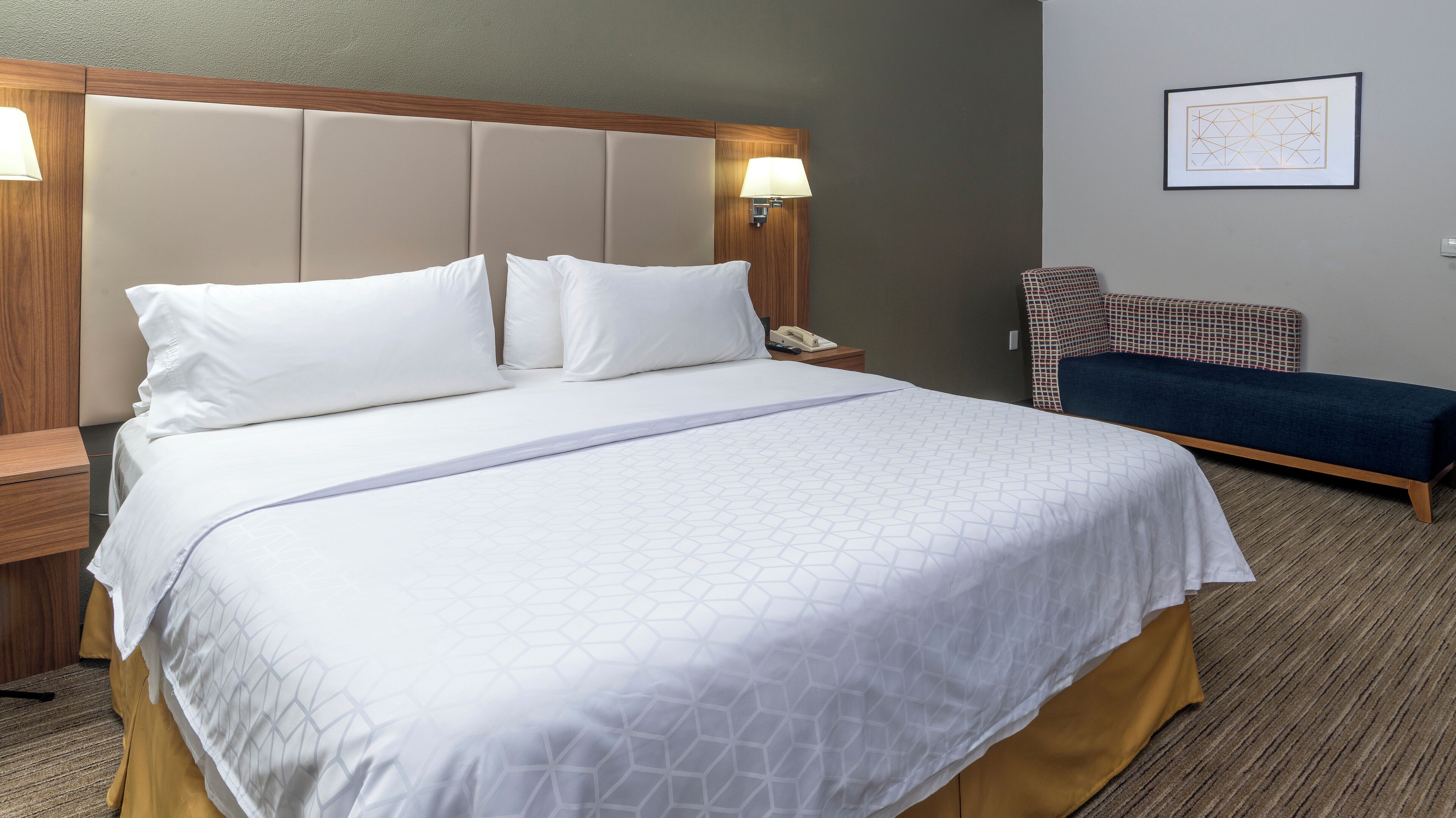 A hotel room with a large bed, a bedside table, a nightstand, a lamp, a telephone, a bench, and a framed picture on the wall.
