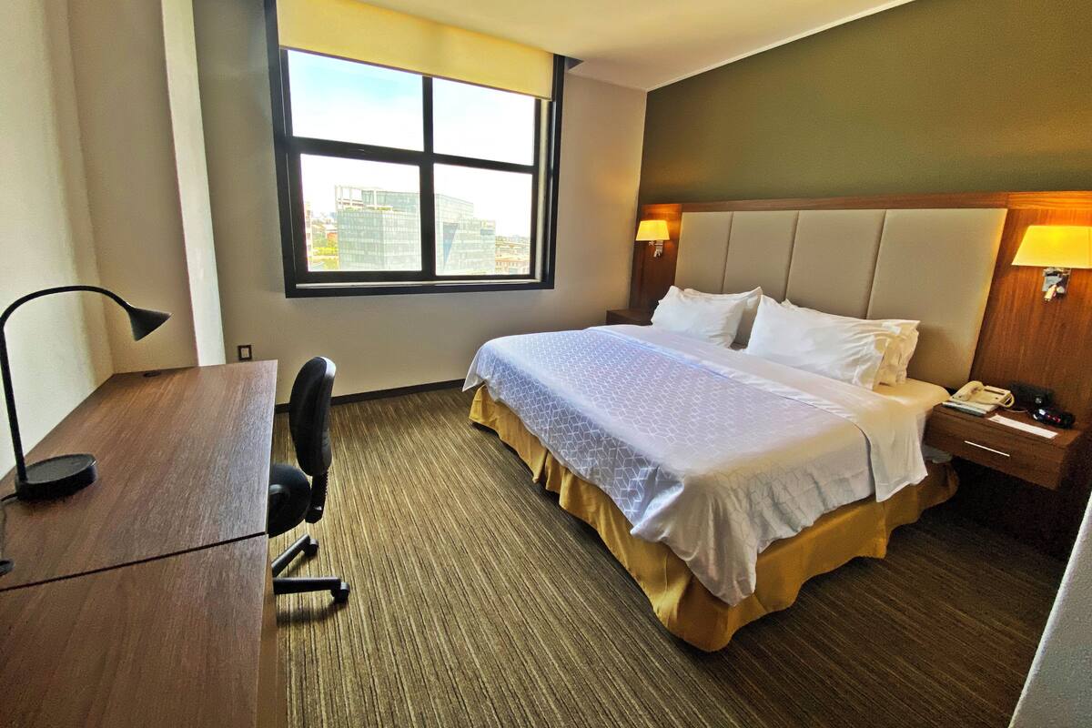 A hotel room with a bed, a desk, a chair, a window with a city view, and a nightstand with a lamp.