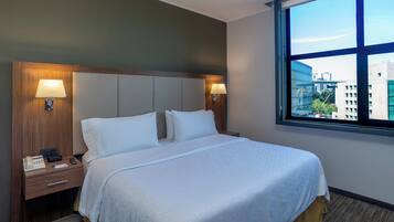 A hotel room with a large bed, two bedside lamps, a nightstand with a phone and a remote control, and a window with a city view.