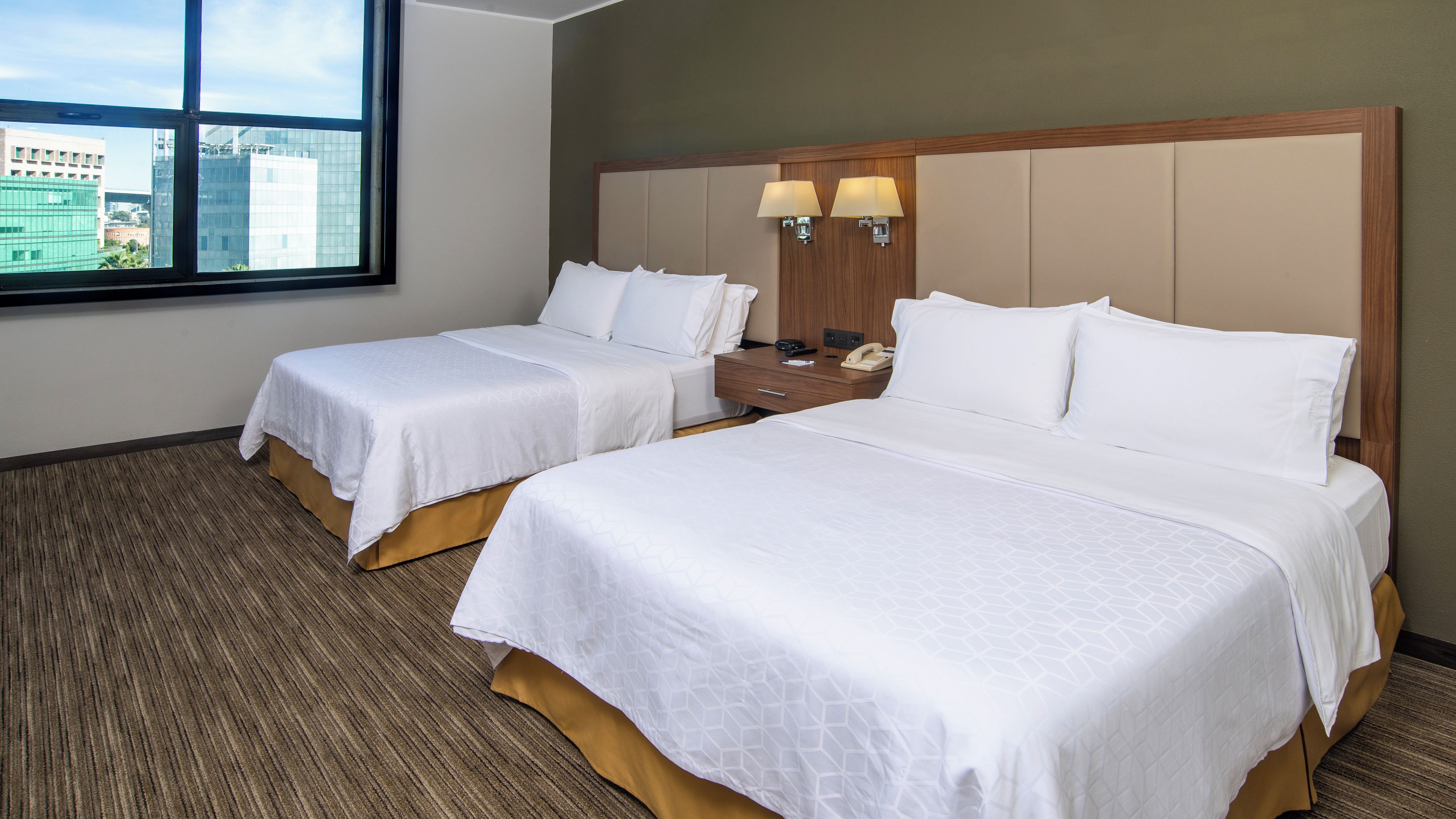 A hotel room with two beds, a nightstand, a lamp, and a window with a city view.