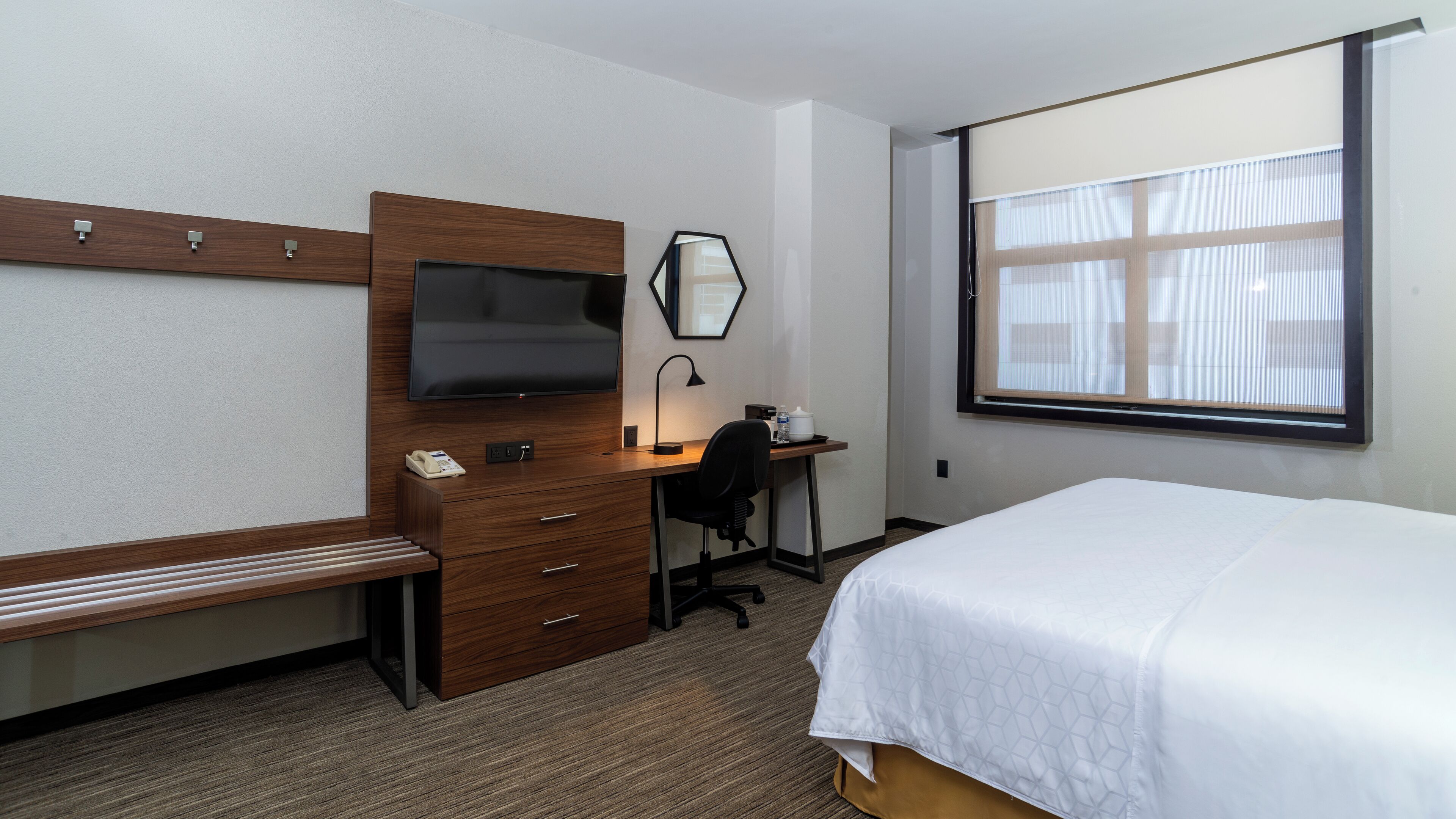 A hotel room with a bed, a desk with a chair, a television, a mirror, and a window.