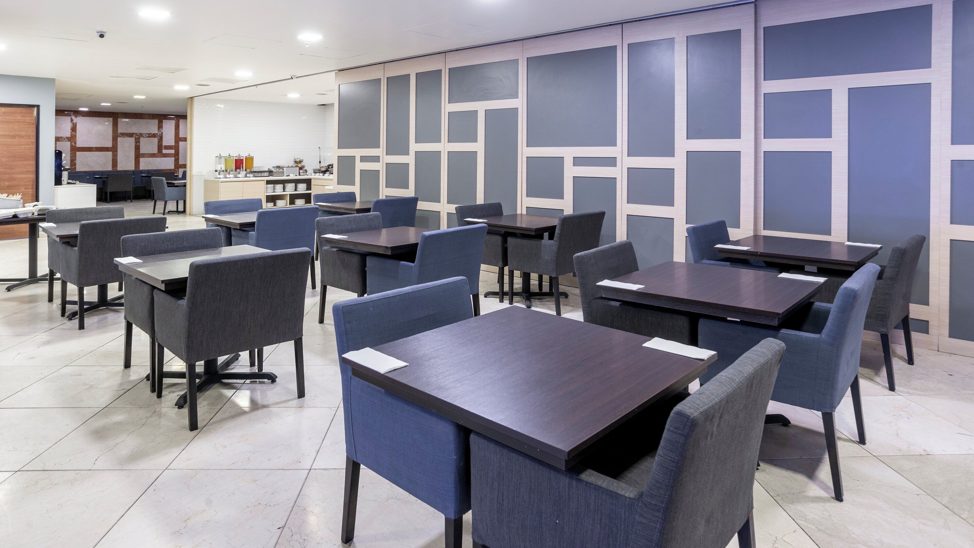 A modern dining area with multiple tables and chairs, a kitchen in the background, and a patterned wall design.