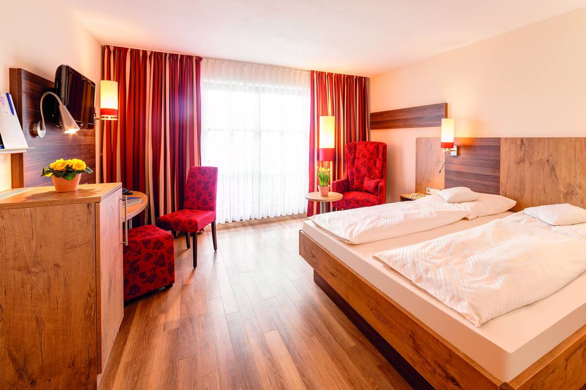 A hotel room with a wooden bed, a red chair, a desk with a lamp, and a TV.