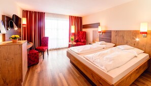A hotel room with a wooden bed, a red chair, a desk with a lamp, and a TV. - Seehotel Wassermann (Seeon-Seebruck)