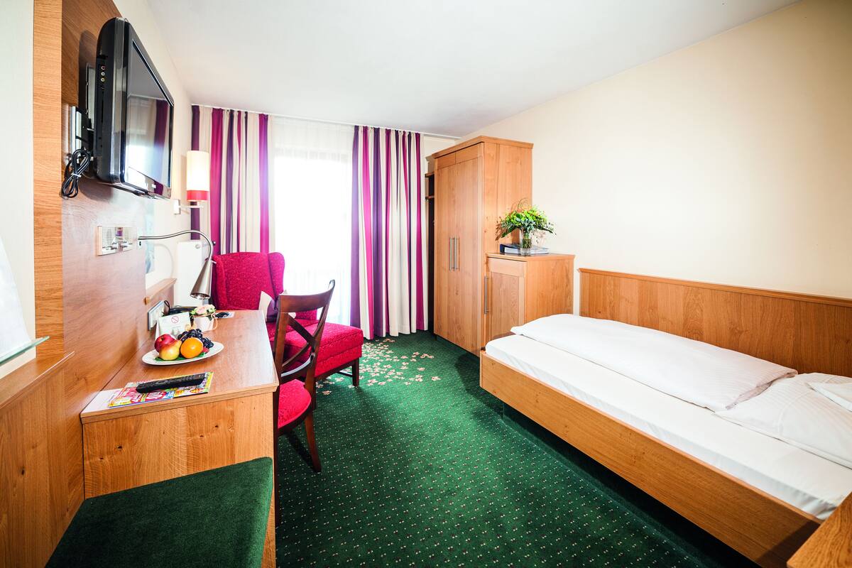 A hotel room with a bed, a desk with a chair, a television, a wardrobe, and a window with curtains.