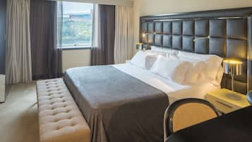 A hotel room with a large bed, a bench, a desk, and a view of the outdoors through the window.