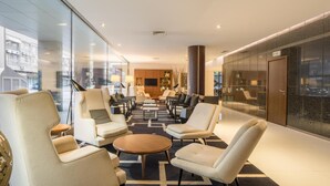 A modern hotel lobby with a central seating area, a reception desk, and a large window offering a city view. - NH Coimbra Dona Inês (Coimbra)