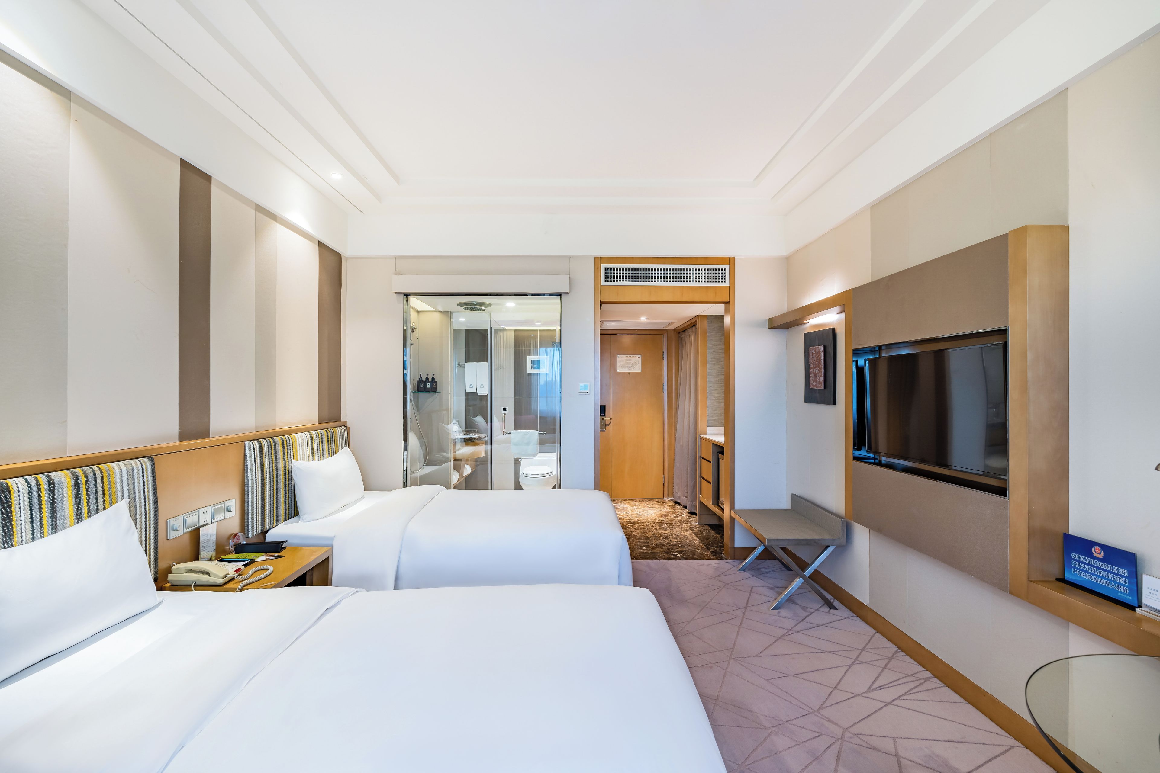 A hotel room with two beds, a TV, a desk, and a bathroom visible through a glass partition.