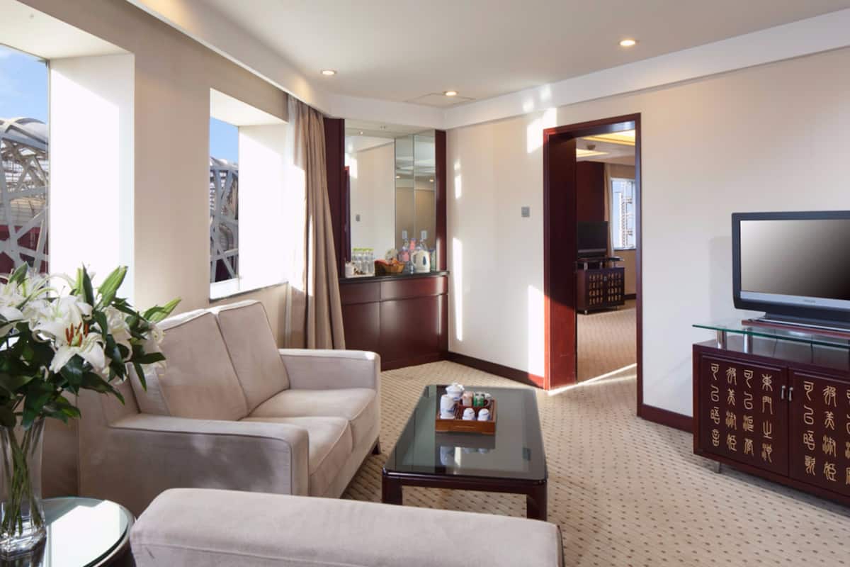 A modern hotel room with a sofa, a glass coffee table, a TV on a cabinet, and a large window with curtains.
