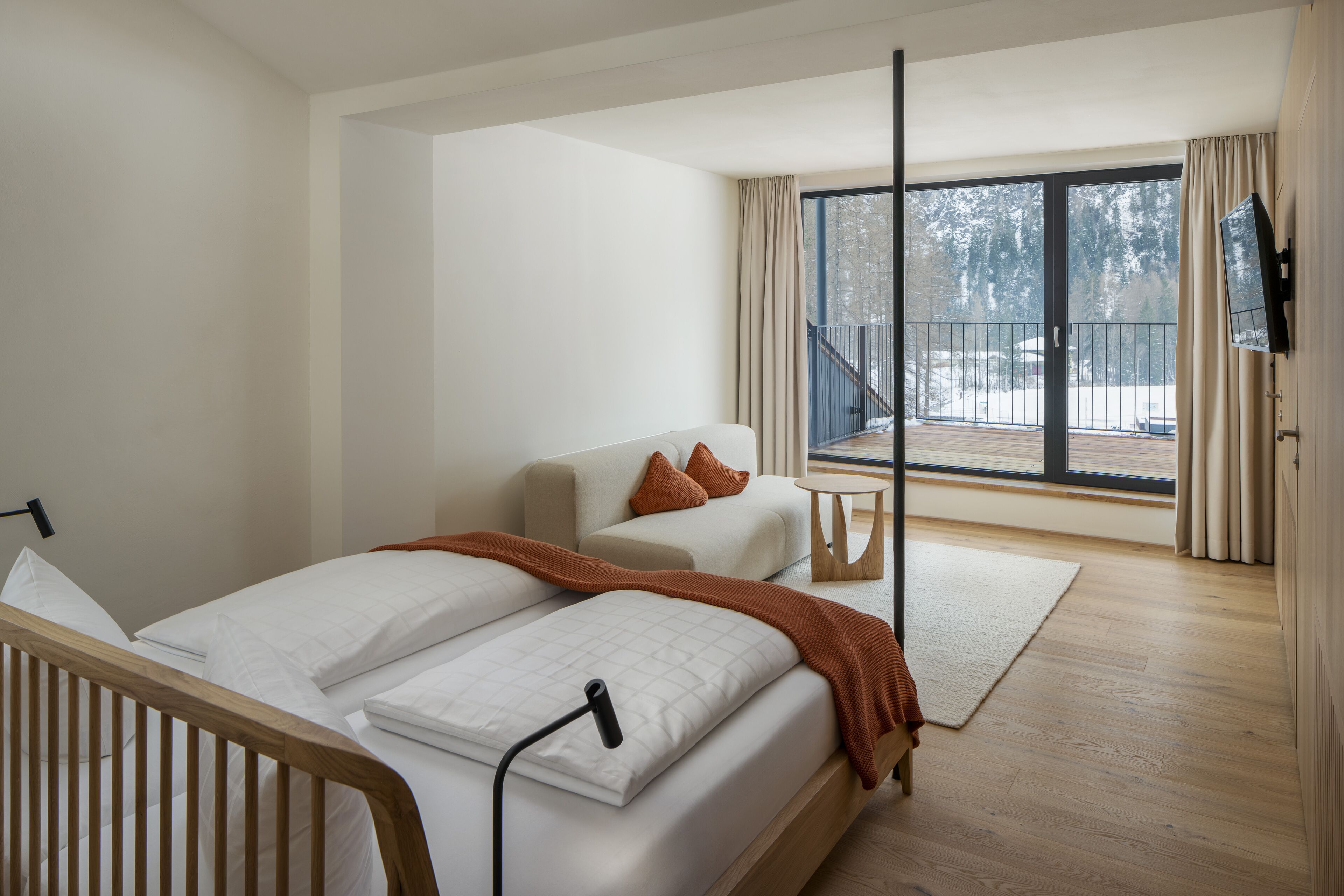 A modern bedroom with a bed, a sofa, a TV, and a balcony with a view of trees.