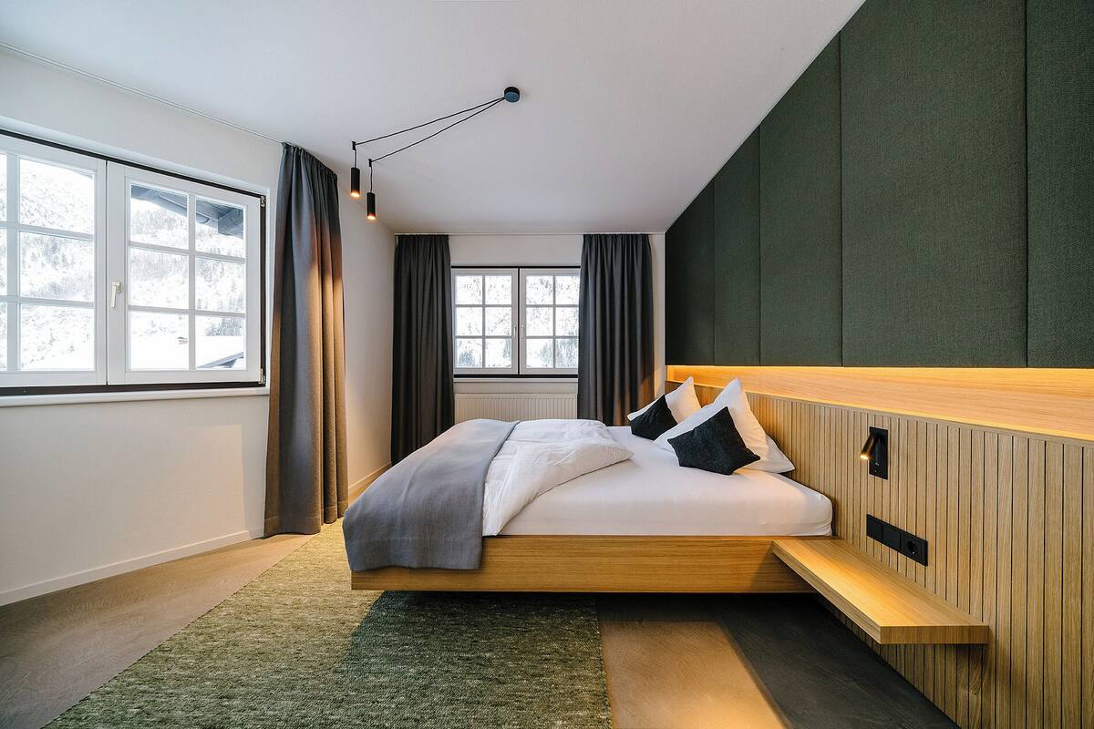 A modern bedroom with a wooden bed frame, a built-in bench, and a view of snowy trees through the windows.