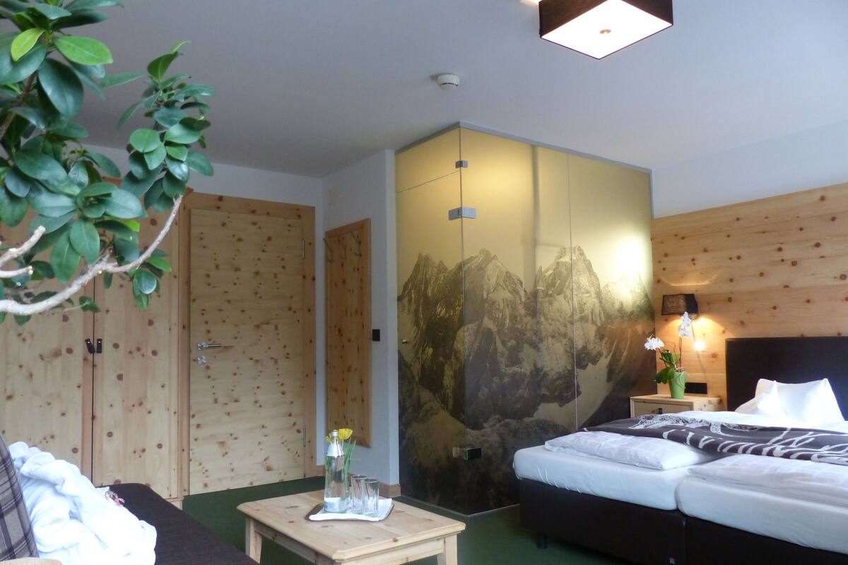 A hotel room with a bed, a small table with a vase, a wall with a mountain painting, and wooden doors.