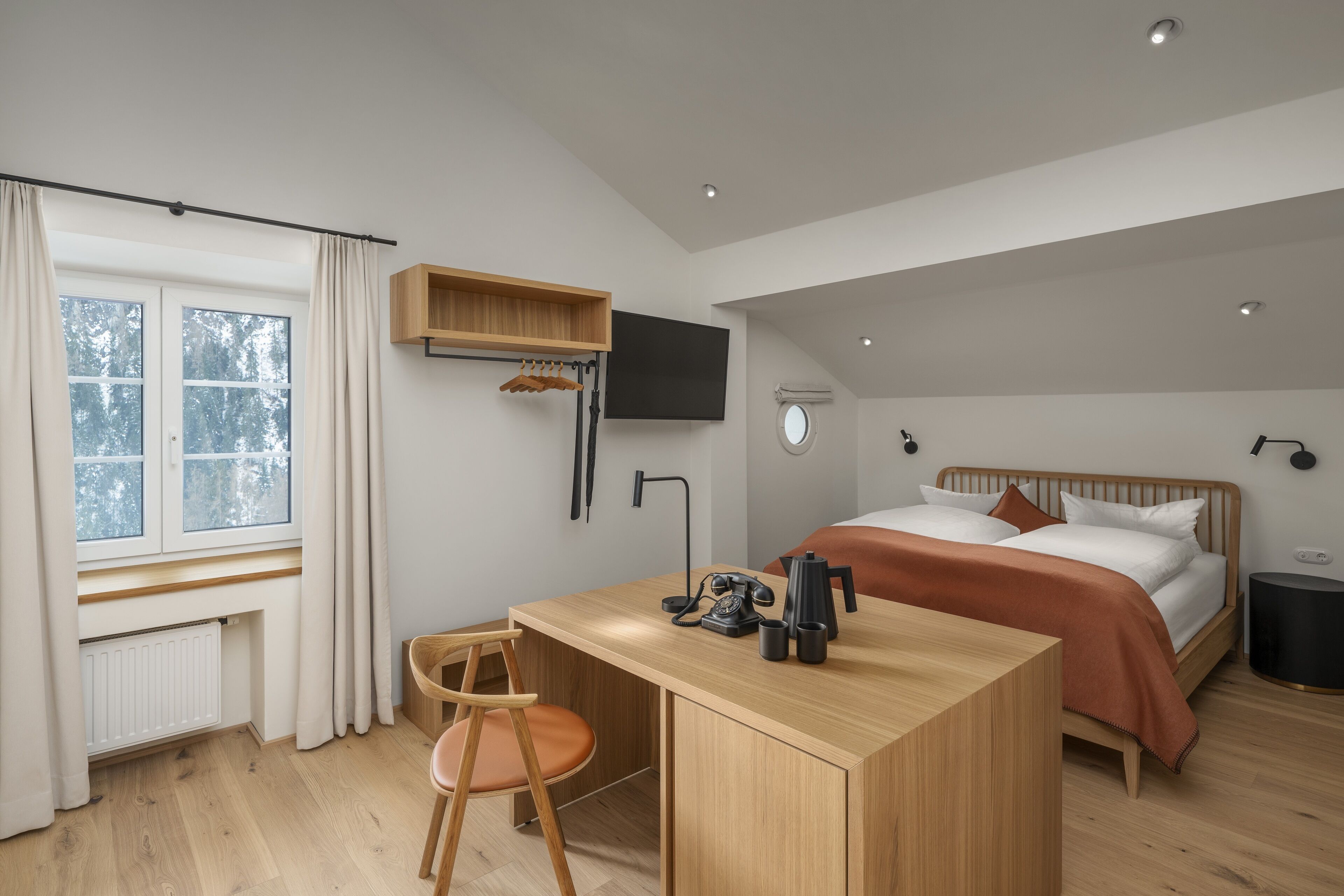 A modern hotel room with a wooden desk, a bed with a brown headboard, a mounted TV, and a window with curtains.