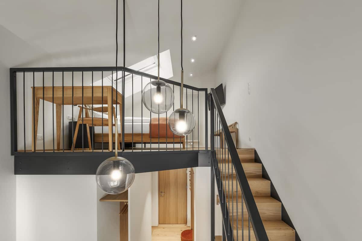 A modern interior with a wooden staircase, a mezzanine level with a bed, and pendant lighting.