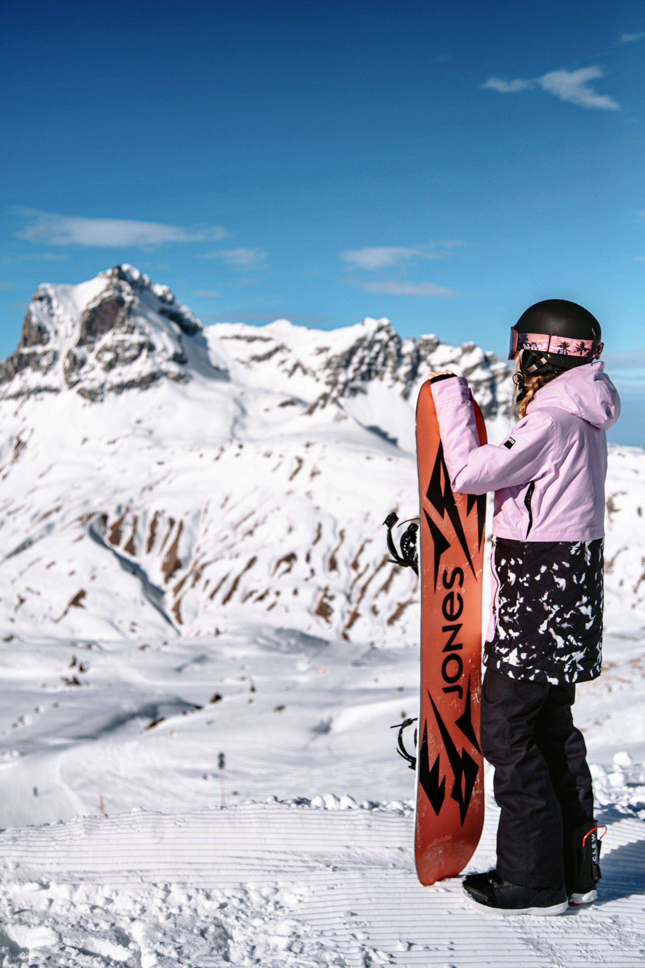 A snowboarder in a pink jacket and black pants holding a snowboard with a black design, standing on a snowy slope with mountains in the background.
