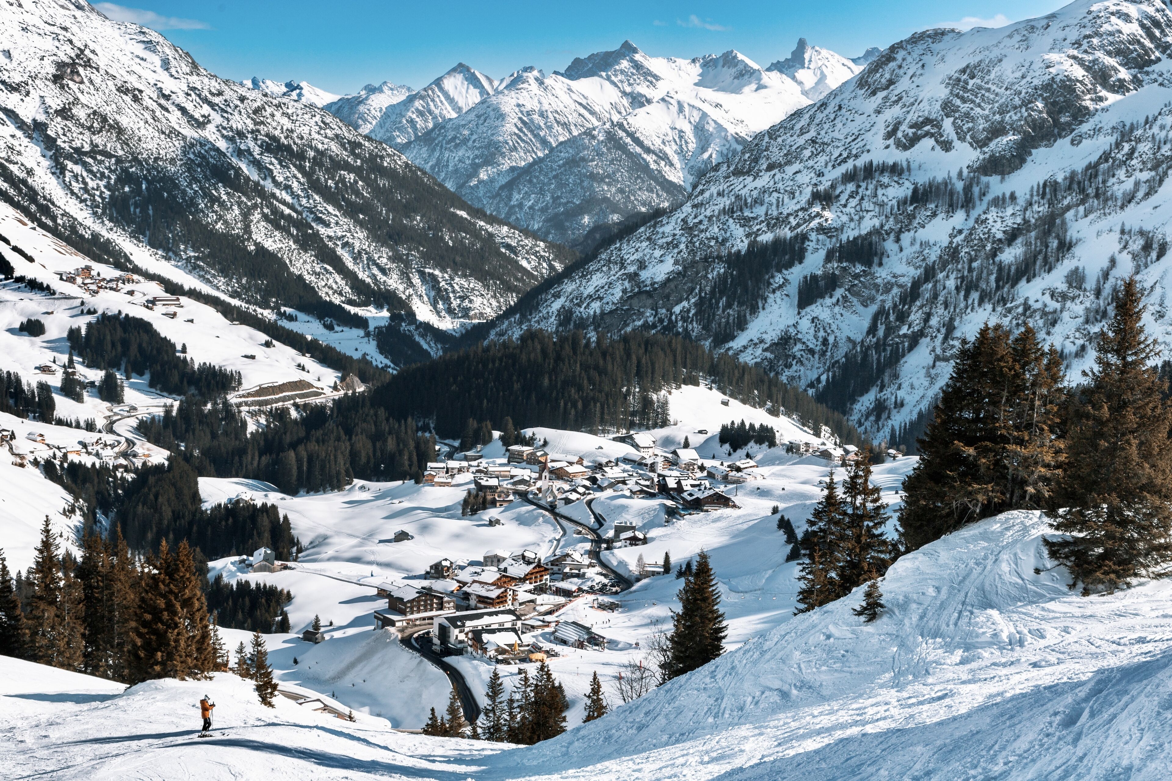 Un village de montagne enneigé, avec des bâtiments blottis dans une vallée, entouré de pentes boisées et de sommets enneigés.