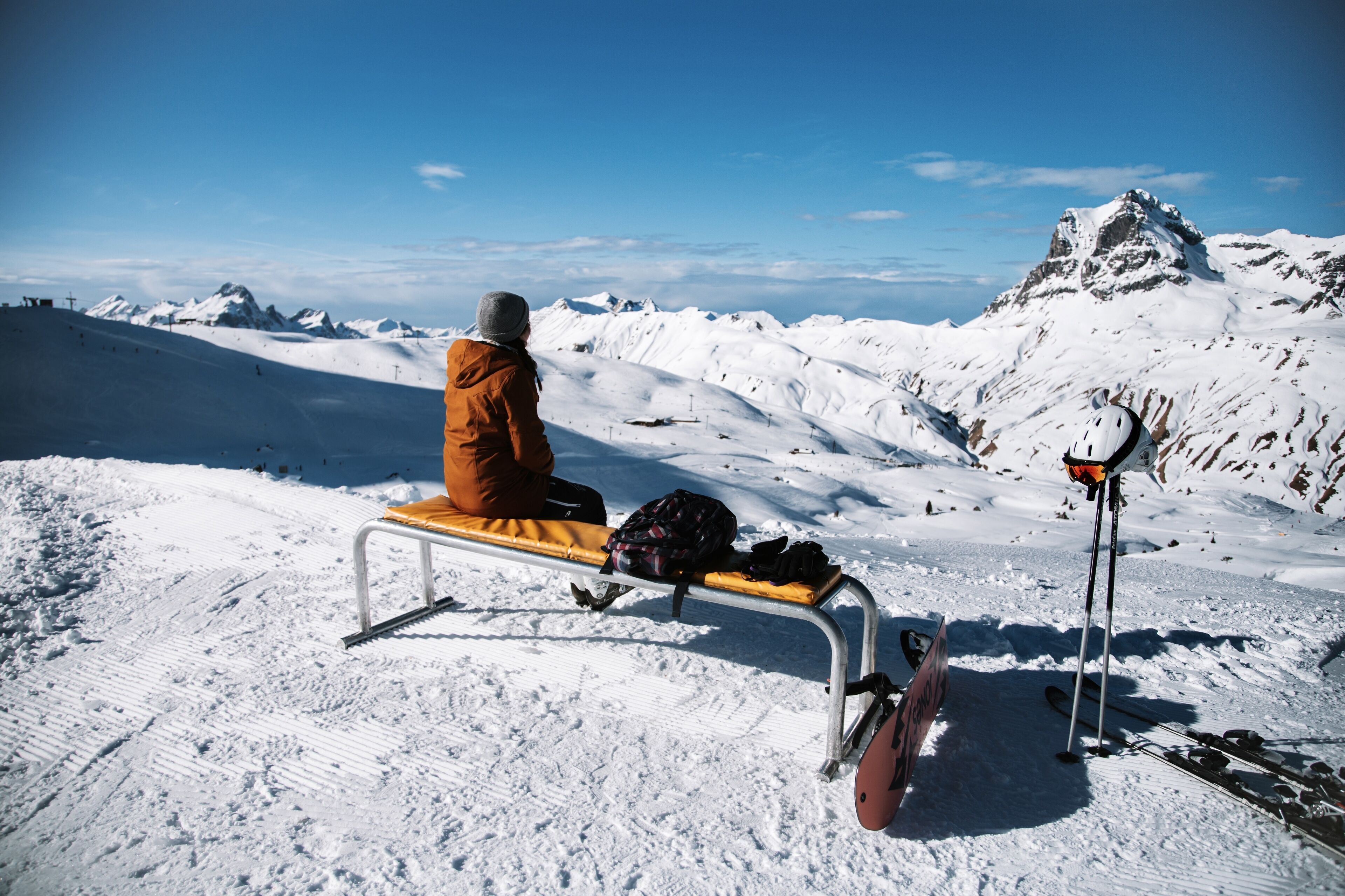 A person sitting on a bench with a backpack and skis leaning against a metal pole, overlooking a snowy mountain landscape.