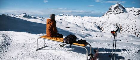 A person sitting on a bench with a backpack and skis leaning against a metal pole, overlooking a snowy mountain landscape.