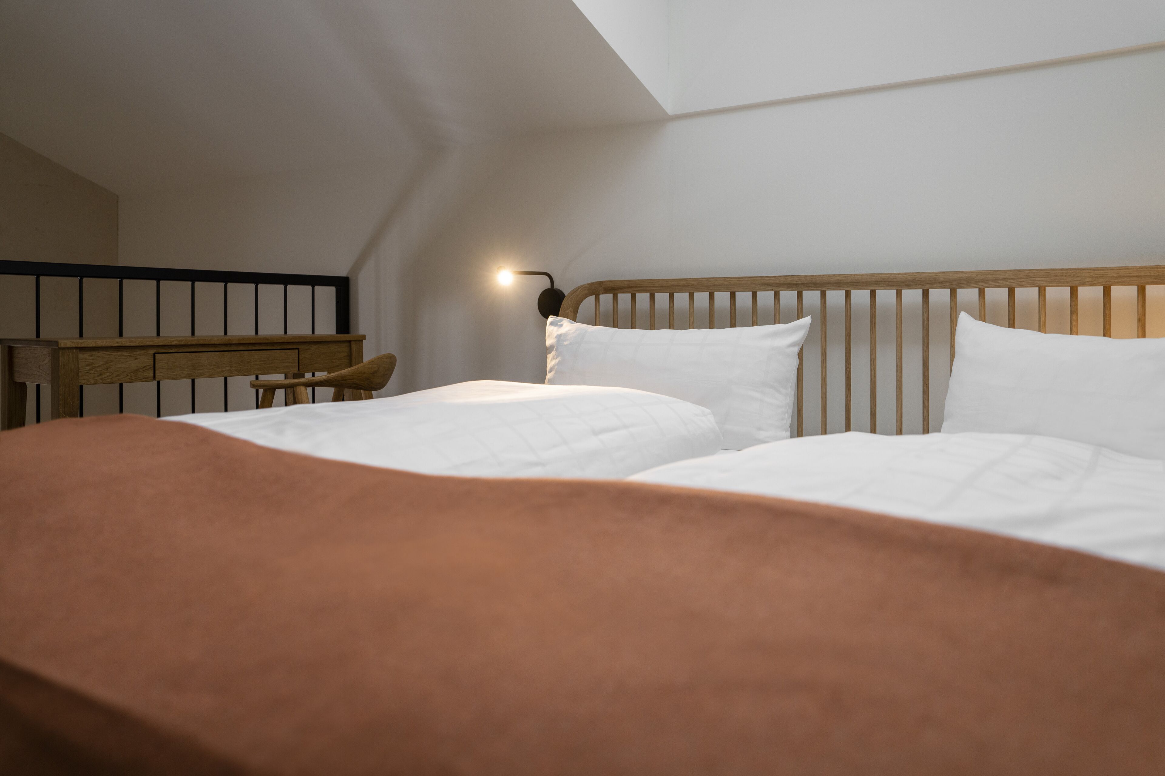 Een slaapkamer met een houten hoofdbord, twee witte bedden, een bruine deken en een nachtkastje met een lamp.