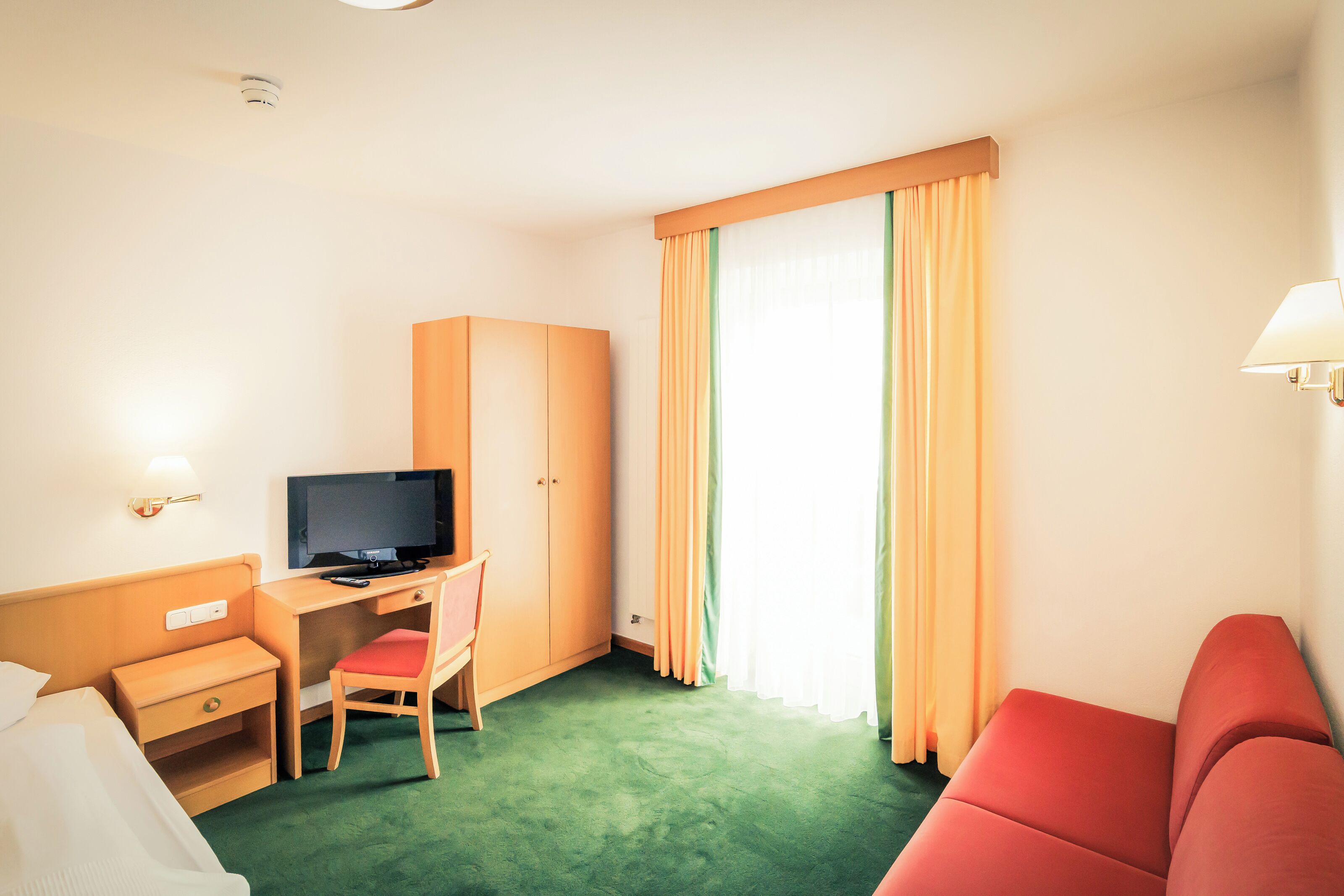 Una habitación de hotel con una cama, un escritorio con televisor, un sofá rojo y una ventana con cortinas.