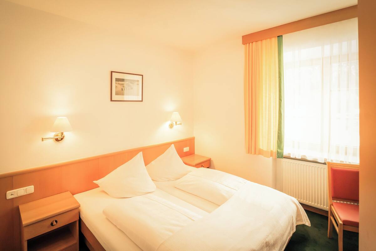 A hotel room with a bed, two bedside lamps, a nightstand, a chair, a window with curtains, and a framed picture on the wall.