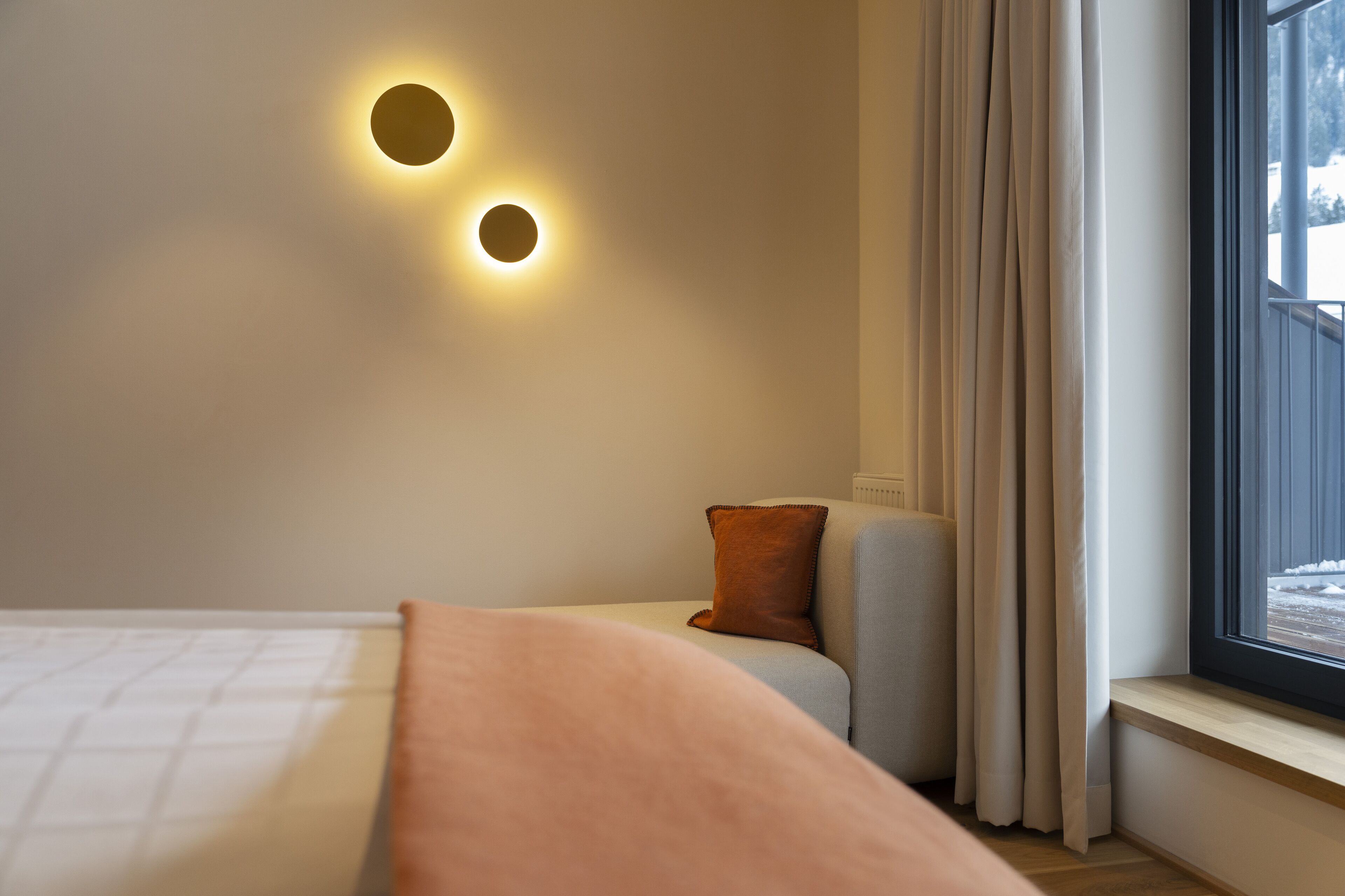 A bedroom with a bed, a nightstand, a window with curtains, and wall-mounted lighting.