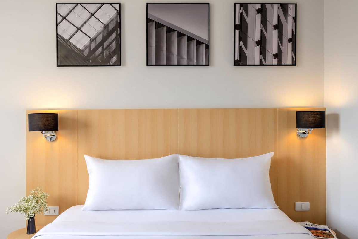 Ein Hotelzimmer mit einem hölzernen Kopfende, zwei Wandlampen, einem Bett mit weißer Bettwäsche und drei gerahmten Bildern an der Wand.