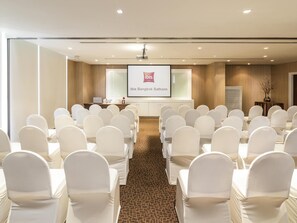 A conference room with rows of white chairs, a podium, a projector screen, and a carpeted floor. - ibis Bangkok Sathorn (Bangkok)