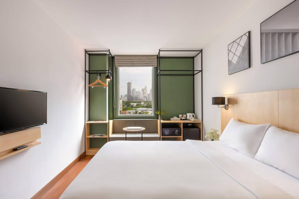A hotel room with a bed, a TV, a window with a city view, a small table, and a shelf with a kettle and phone.
