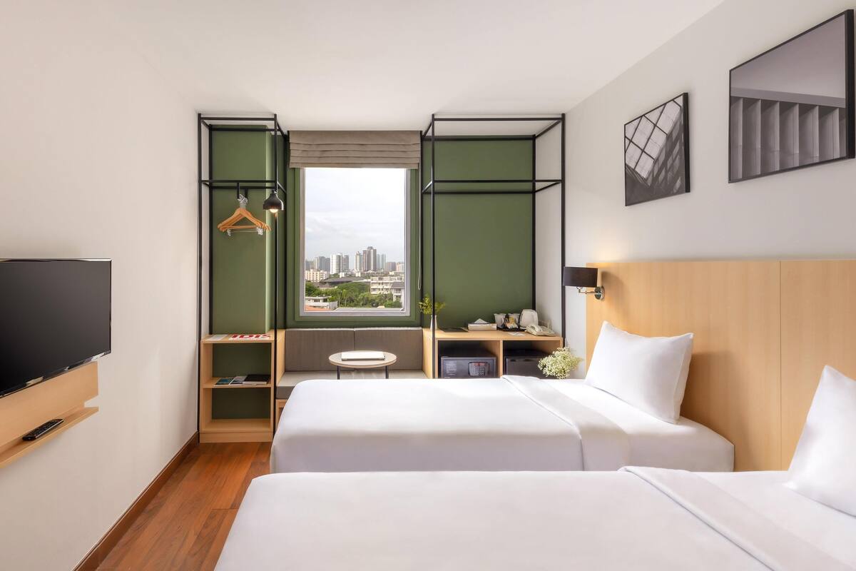 A hotel room with two beds, a TV, a desk with a chair, a window with a city view, and a hanging light fixture.