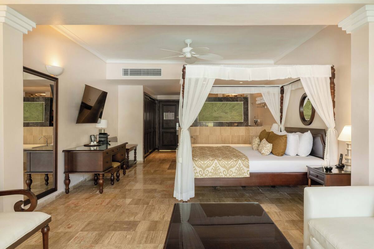 A bedroom with a four-poster bed, a desk with a chair, a television, and a bathroom visible in the background.