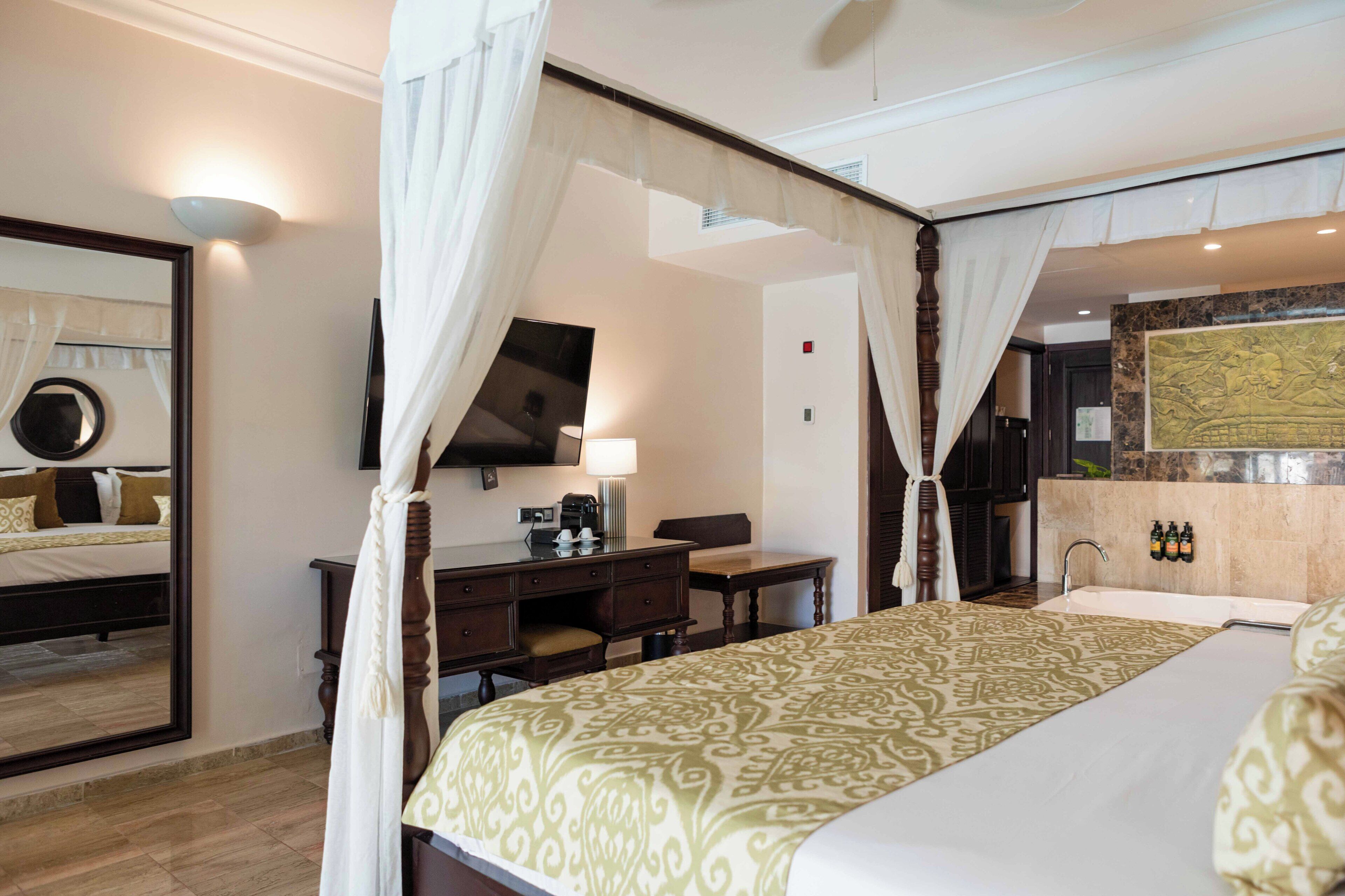 A hotel room with a four-poster bed, a desk with a chair, a television, and a bathroom with a sink and mirror.