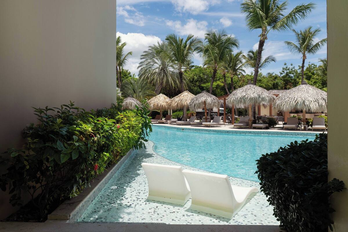 A pool with lounge chairs and thatched umbrellas, surrounded by palm trees.