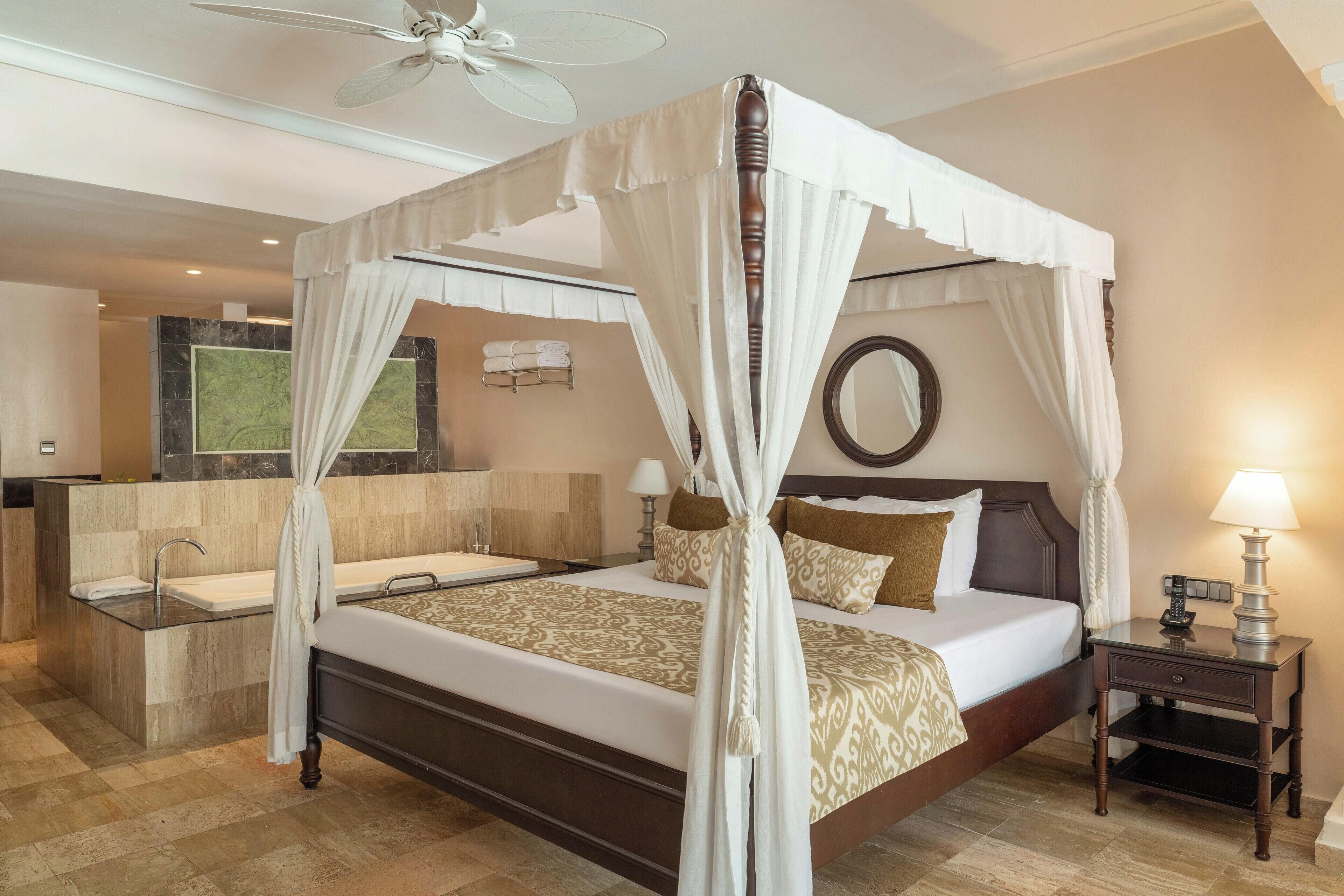 A four-poster canopy bed with a bedside table and lamp, a separate bathroom with a bathtub, and a large mirror on the wall.