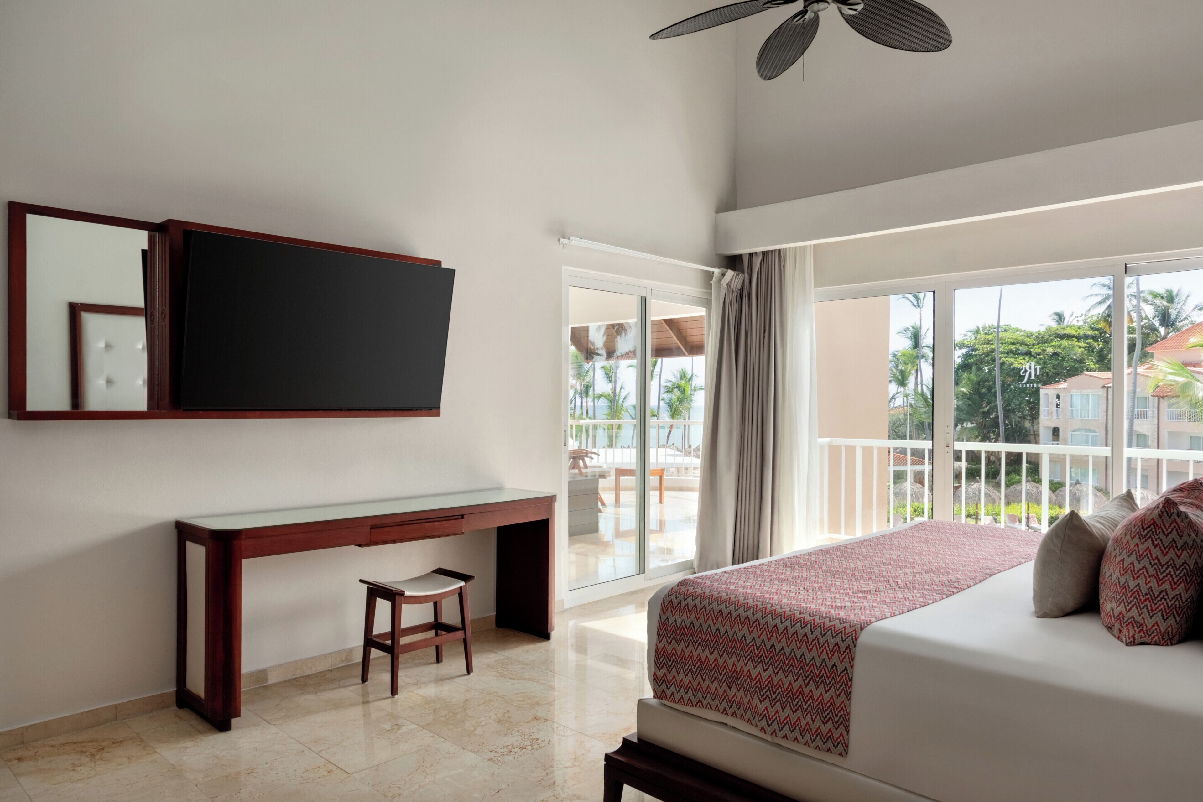 Habitación de hotel con cama, escritorio, silla, televisión y balcón con vistas al exterior.
