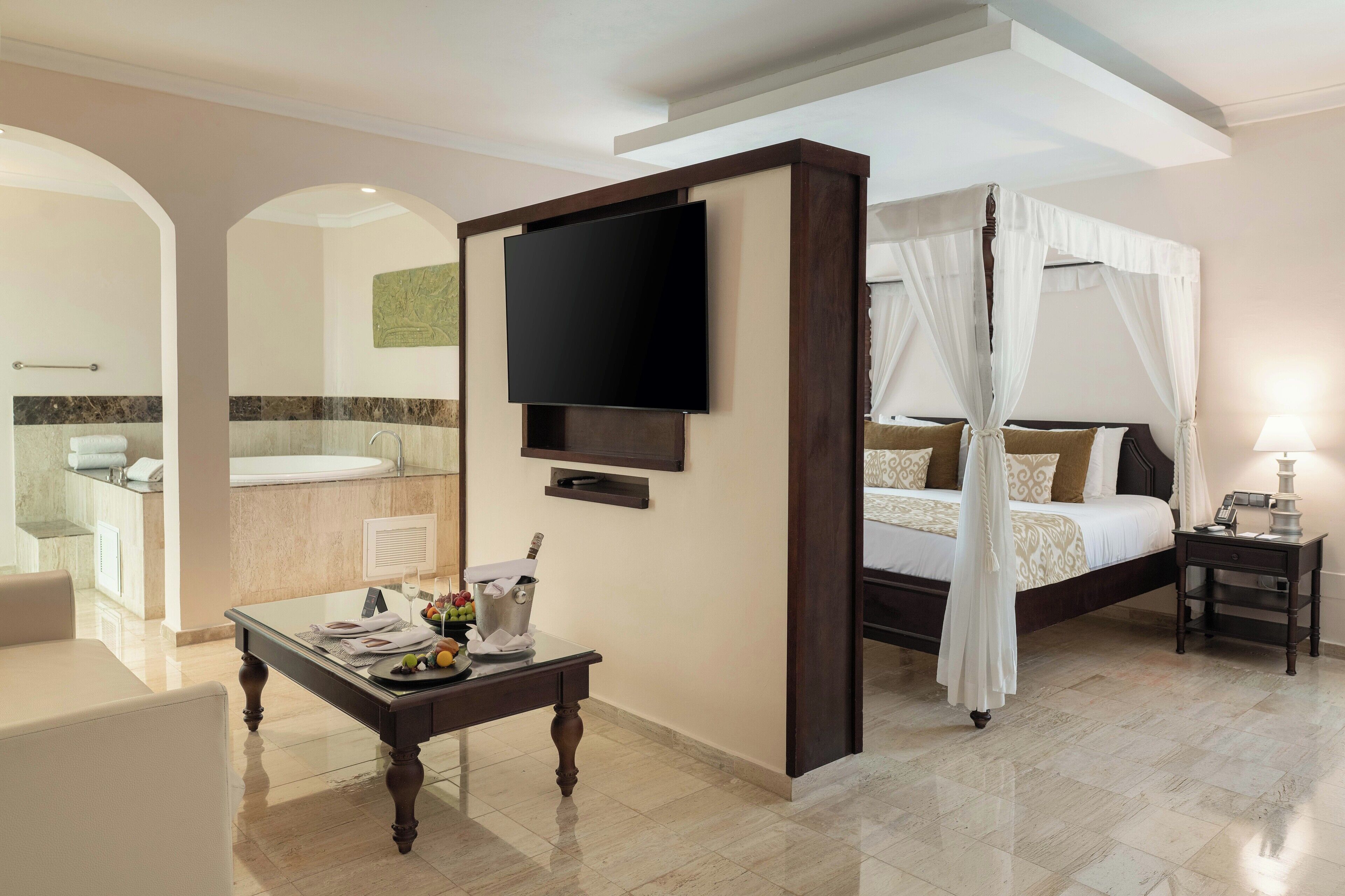 Habitación de hotel con cama, televisor, mesita de noche, una pequeña mesa con una bebida y fruta, y un baño visible a través de un arco.