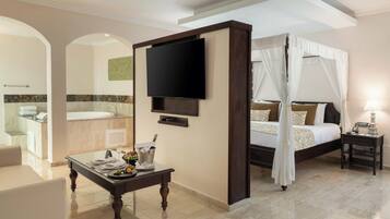 Habitación de hotel con cama, televisor, mesita de noche, una pequeña mesa con una bebida y fruta, y un baño visible a través de un arco.