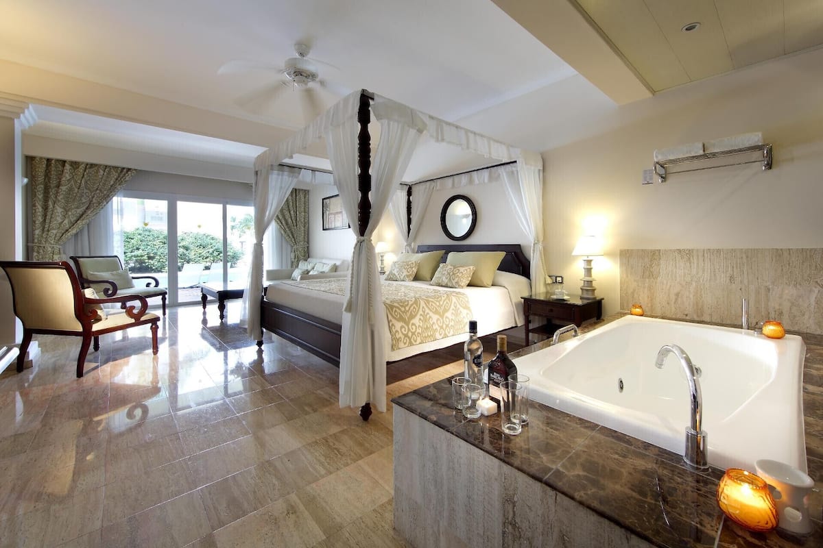 A bedroom with a large bed, a sitting area, a bathroom with a bathtub, and a view of the outdoors.