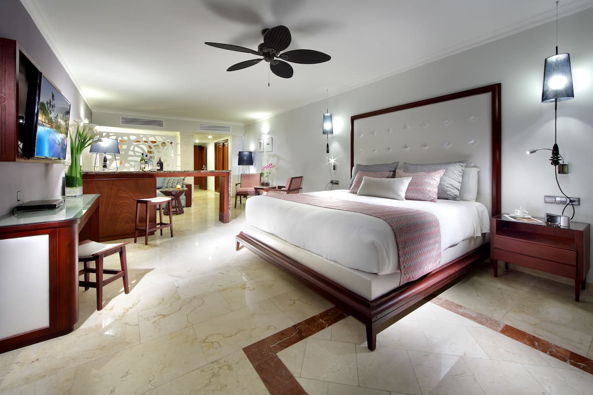 A spacious hotel room with a large bed, a desk, a chair, and a ceiling fan.