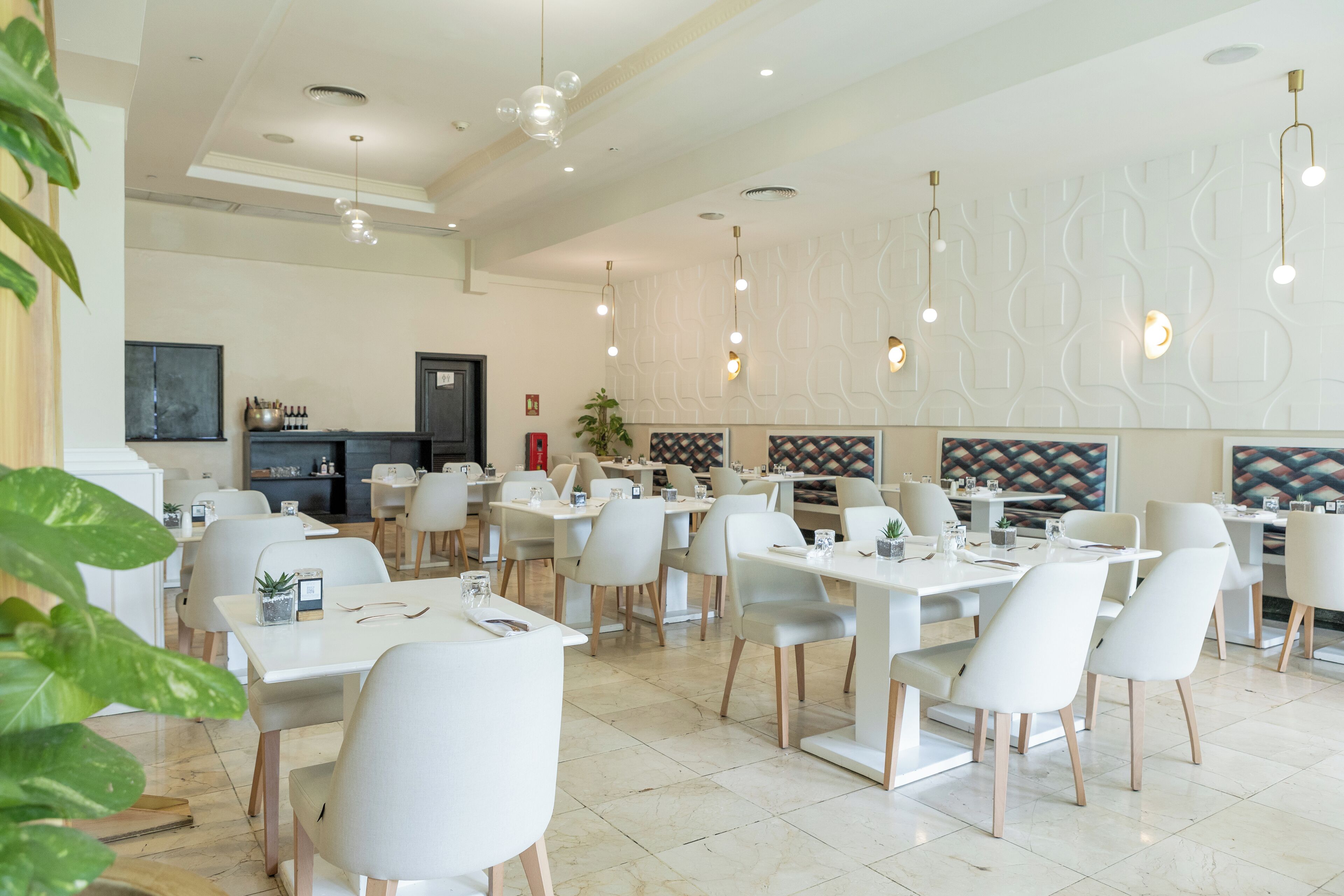 Un restaurante moderno con mesas y sillas blancas, iluminación colgante y un diseño de pared estampado.