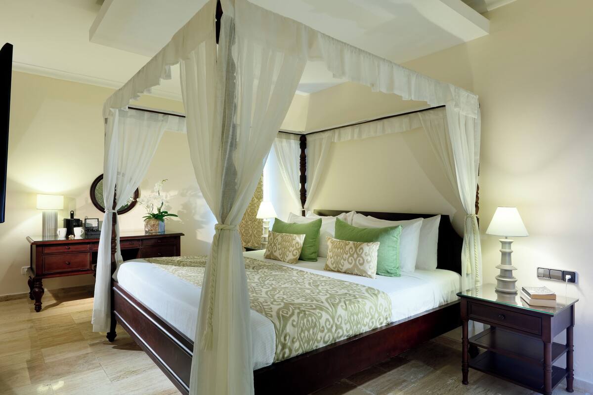 A four-poster canopy bed with a bedside table and lamp, a nightstand with a lamp, and a bedside plant.