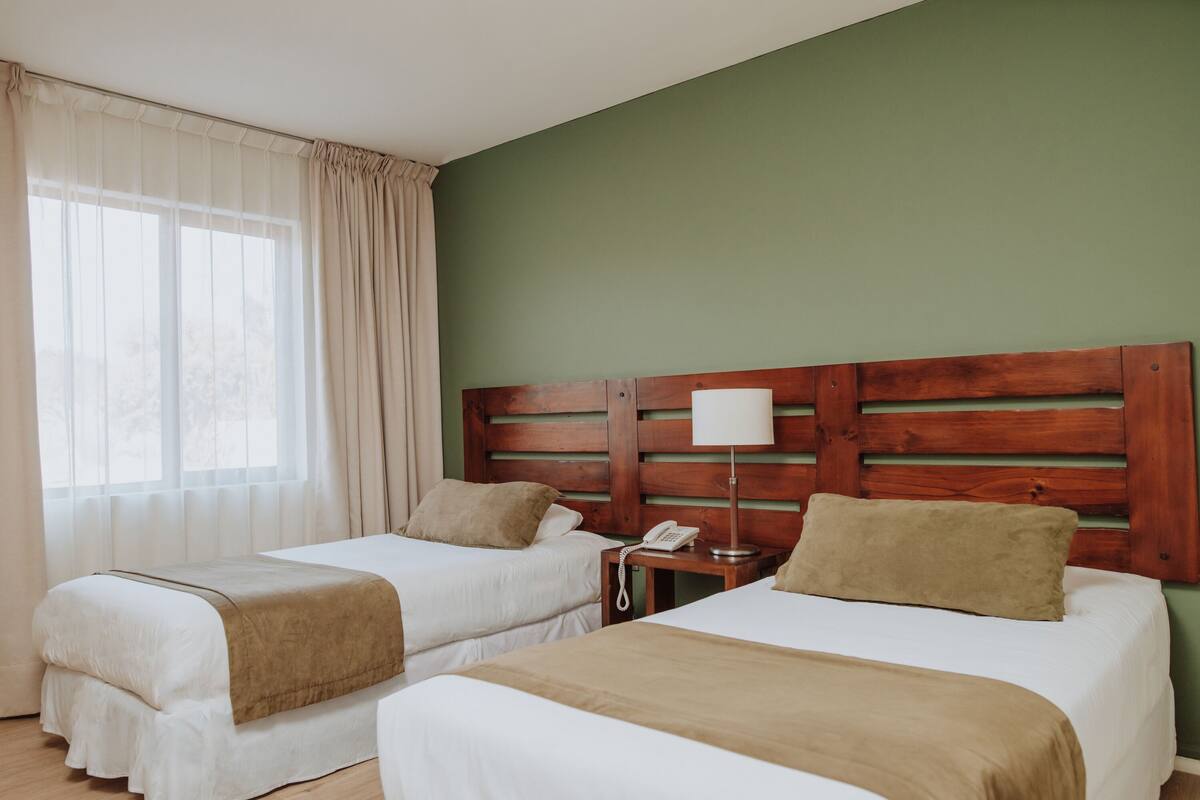 Ein Hotelzimmer mit zwei Betten, einem hölzernen Kopfteil, einem Nachttisch mit Lampe und einem Fenster mit Vorhängen.