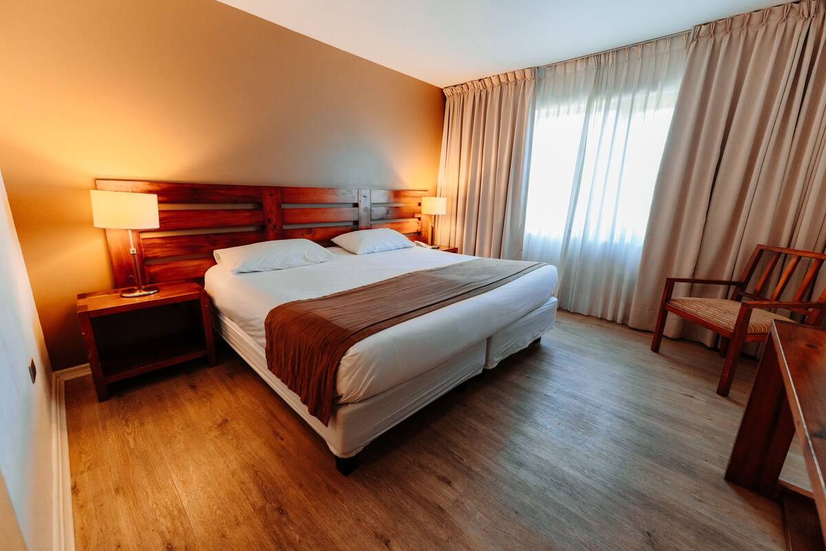 Ein Hotelzimmer mit einem großen Bett, zwei Nachttischen mit Lampen, einem hölzernen Kopfteil, einem Sessel und einem großen Fenster mit Vorhängen.