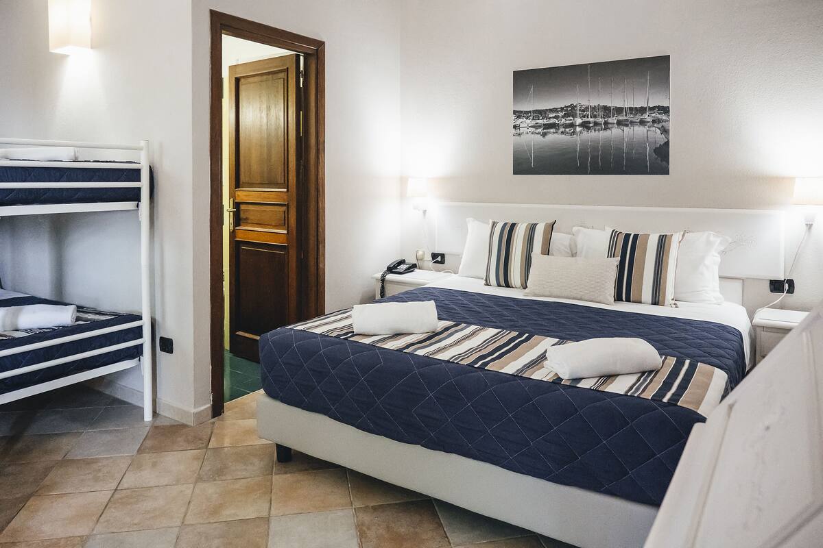Une chambre d’hôtel avec un lit superposé, un grand lit avec une literie à rayures, une table de chevet, une lampe et un tableau accroché au mur.