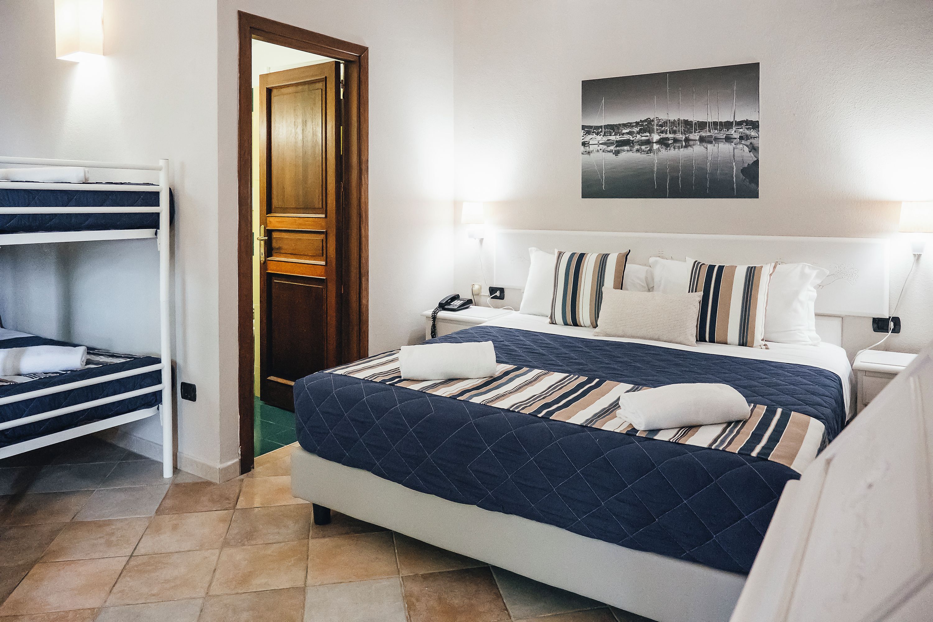 Ein Hotelzimmer mit einem Etagenbett, einem großen Bett mit gestreifter Bettwäsche, einem Nachttisch, einer Lampe und einem an der Wand hängenden Bild.