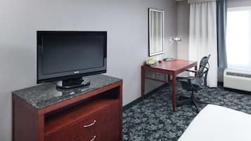 Una habitación de hotel con televisor de pantalla plana sobre un soporte de madera, un escritorio con silla, una cama y un ventanal con cortinas.
