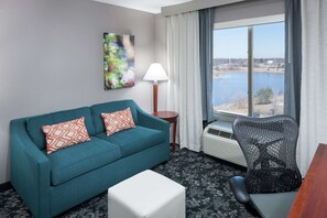 A teal sofa with patterned pillows, a white ottoman, a black office chair, a lamp, and a view of a body of water through a window. - Hilton Garden Inn Naperville/Warrenville (Warrenville)