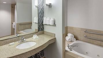 A modern bathroom with a granite countertop sink, a large mirror, a bathtub with grab bars, and a towel rack with towels.