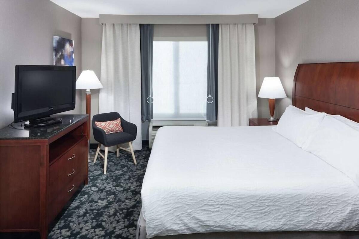 Une chambre d’hôtel avec un lit, un bureau avec une télévision, deux lampes, une chaise et une fenêtre avec des rideaux.